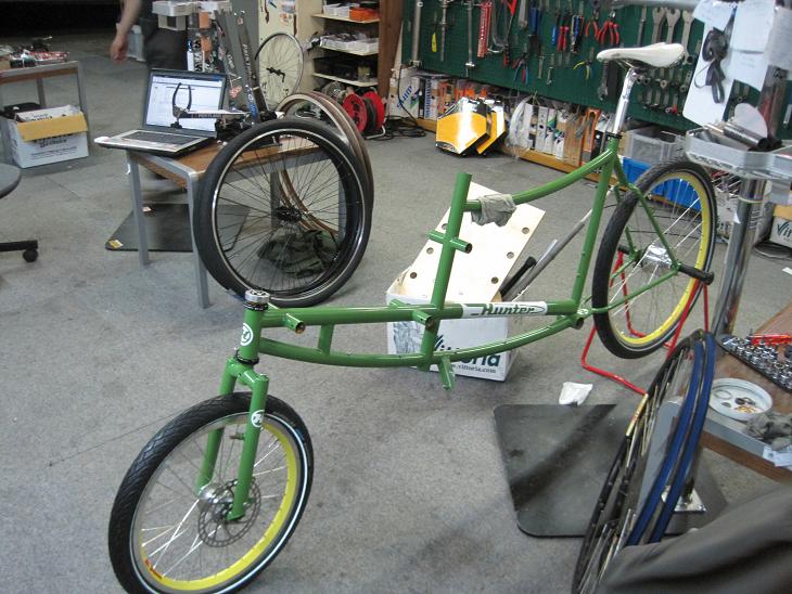 Front, left side view of a green 2 person bike, being assembled, inside of a bike shop